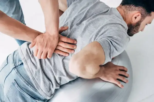 Terapeuta wykonuje masaż pleców mężczyzny na piłce rehabilitacyjnej. Czy osteopata jest lekarzem? To pytanie nurtuje wielu pacjentów.
