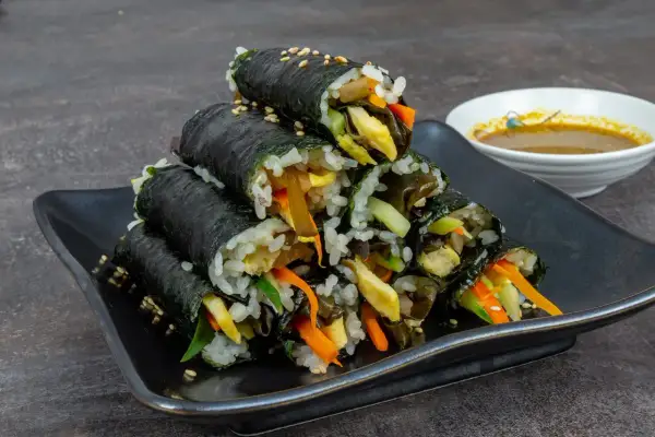 Gestapeltes Kimbap, koreanische Reisrollen mit Gemüse und Ei, auf schwarzer Platte, daneben eine Schale Dip.