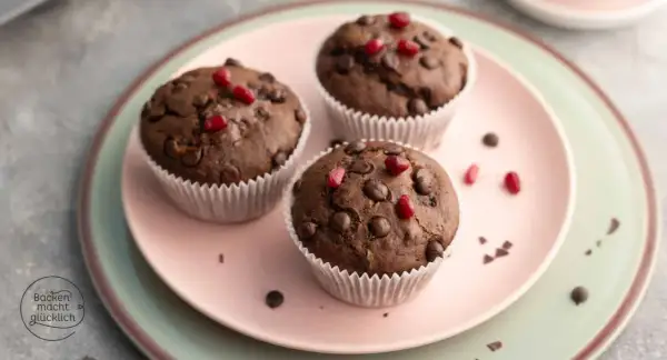 Drei saftige, vegane Muffins mit Schokostückchen und Granatapfelkernen auf einem rosa Teller.