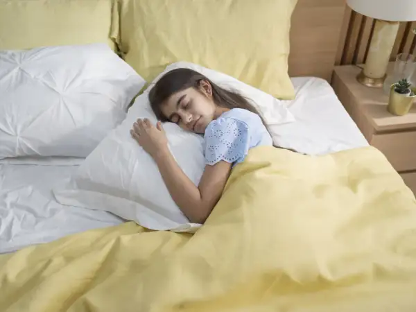 A young girl sleeps peacefully under a yellow blanket. This serene scene reminds us of the importance of fresh air and how to get rid of off-gassing smells for a healthy sleep environment.