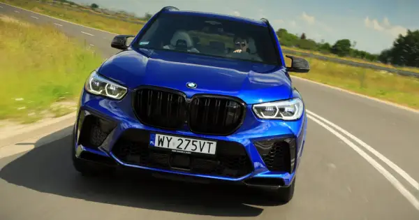 Niebieski BMW X5 M Competition pędzi po drodze, kierowca w okularach przeciwsłonecznych za kierownicą.