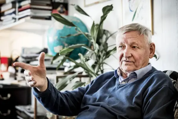 Äldre man med grått hår, liknar Ragnar Skanåker, gestikulerar med handen. Bakom honom syns en jordglob och växter.