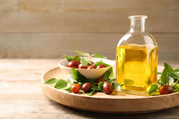 Flasche mit goldenem Simmondsia Chinensis Seed Oil neben einer Schale mit Jojobasamen und grünen Blättern auf einem Holzteller.
