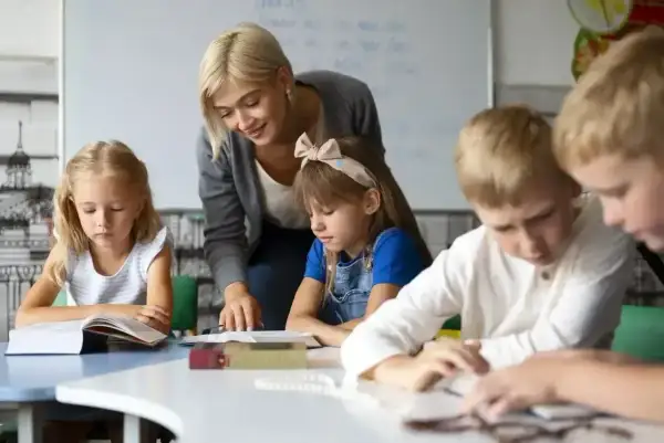 Nauczycielka wspiera uczniów w nauce, pokazując swoje kompetencje rady pedagogicznej. Dzieci skupione na książkach.