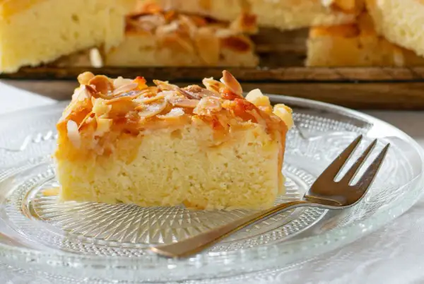 Ein Stück saftiger **Mandelkuchen vom Blech** mit knusprigen Mandelblättchen und Zucker bestreut, auf einem Glas-Teller mit einer Gabel.