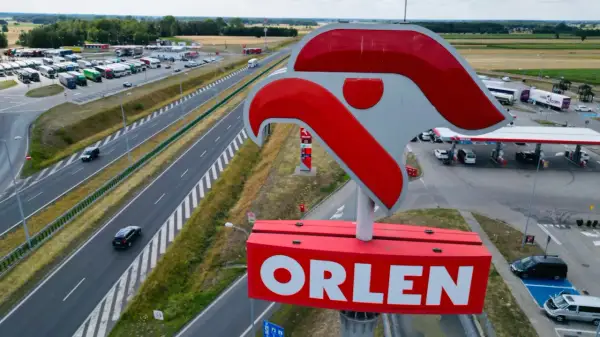 Duży znak ORLEN nad stacją paliw przy autostradzie. W tle parking z ciężarówkami i samochodami.