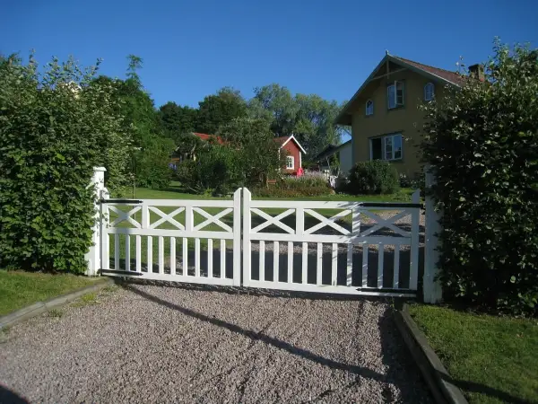 Vit grind med korsmönster leder in på en grusgång entre till ett gult hus och en röd stuga.