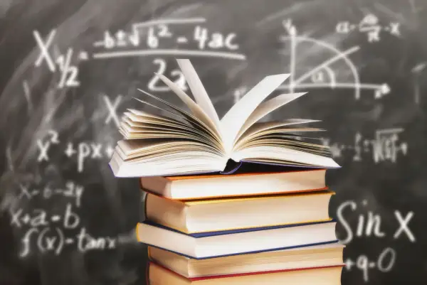 Stos książek z otwartą księgą na wierzchu na tle tablicy z równaniami matematycznymi.