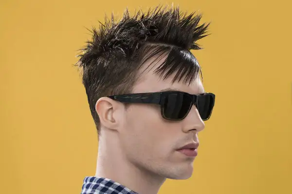 Uomo con capelli a spazzola corti, occhiali da sole scuri e camicia a quadri blu e bianchi su sfondo giallo.