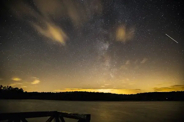 Nocne niebo nad jeziorem, pełne gwiazd i smug chmur, idealne do nauki, jak fotografować gwiazdy. Widać Drogę Mleczną i spadające gwiazdy.