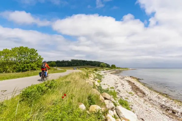 En cyklist njuter av en vacker dag längs en kustväg, en av många cykelleder i Danmark. Havet glittrar bredvid den steniga stranden.