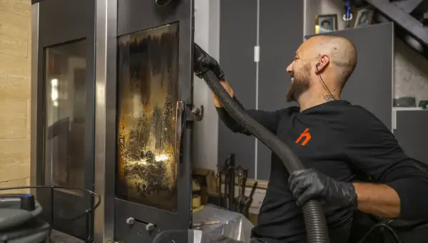 Un homme nettoie la vitre d'un poêle avec un aspirateur. Un bon entretien garantit la bonne fréquence de ramonage.