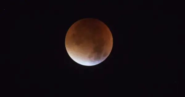 Czerwona poświata na Księżycu podczas zaćmienia. Widoczny fragment tarczy Księżyca w odcieniach pomarańczu i brązu na tle czarnego nieba.