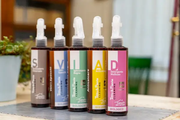Cinque flaconi spray con etichette colorate: "S" (Formulatore Universale), "V" (Vetri Specchi), "I" (Igienizzante Bagno), "A" (Anticalcare), "D" (Detergente Multiuso). La differenza tra detergente e detersivo è chiara: qui sono tutti detergenti biologici.