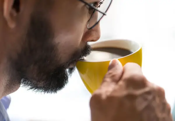Man med glasögon och skägg dricker ur en gul mugg. Kanske känner han yrsel av kaffe.