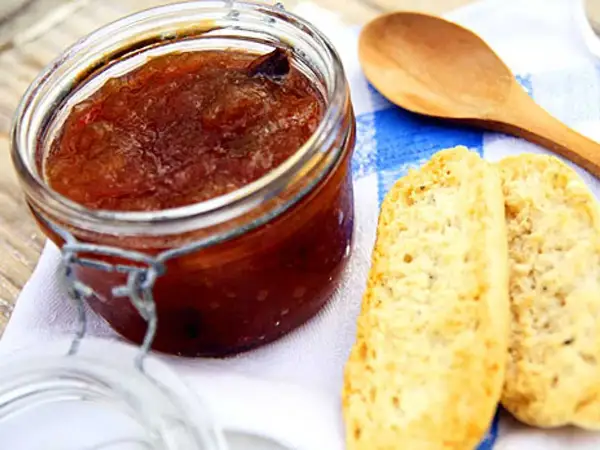 En burk med hemlagad rabarbermarmelad står bredvid två bitar rostat bröd och en träsked på en blåvitrandig duk.