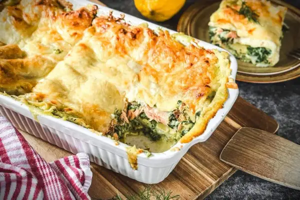 Herzhafte Spinat-Lachs-Lasagne, goldbraun überbacken, dampft in einer weißen Auflaufform. Ein Stück auf einem Teller daneben zeigt die Schichten.