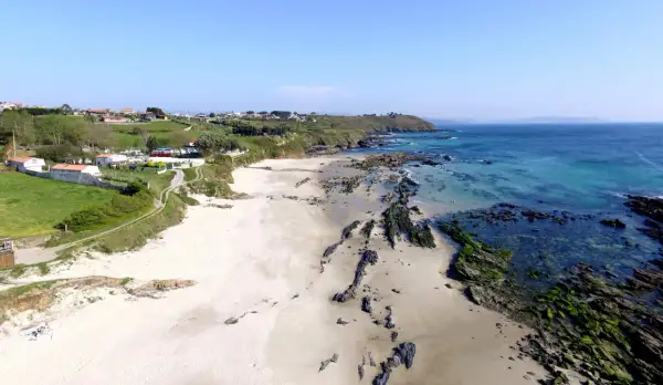 Vista aérea de la playa pragueira, con arena blanca, rocas cubiertas de algas y el mar azul turquesa. Casas y colinas verdes al fondo.