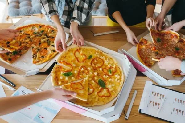 Kilka osób bierze kawałki pizzy z pudełek na drewnianym stole.