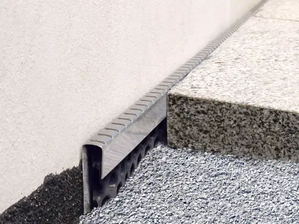 Detailansicht: Noppenbahn einbauen an einer Hauswand mit Granitstufe und Schotterbett.