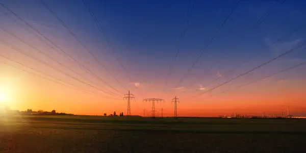 Linie energetyczne biegnące nad polem przy zachodzie słońca. W oddali widać przemysłowy krajobraz, przypominający o opłacie sieciowej stałej.