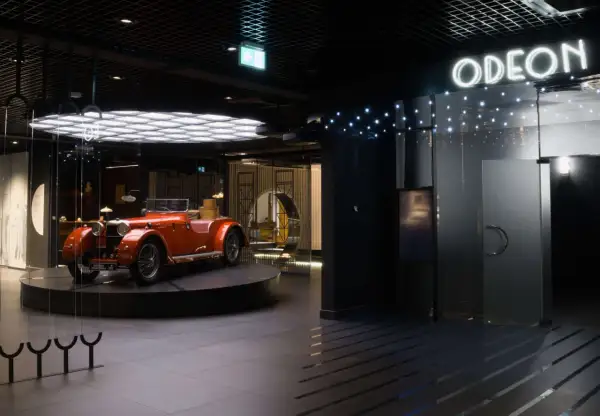 Pomarańczowy kabriolet z lat 20. XX wieku, symbol sztuki dwudziestolecia międzywojennego, prezentowany w nowoczesnym wnętrzu z neonowym napisem "ODEON".