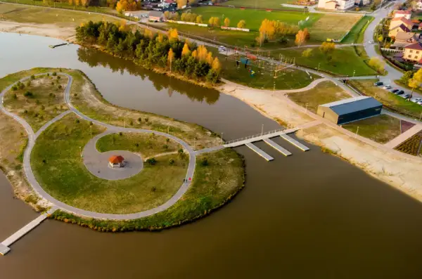 Malownicze, nisko jezioro z wyspą, ścieżkami, altanką i nowoczesnym budynkiem. W tle widać drzewa, plażę i zabudowania.