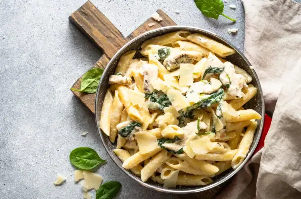 Penne arrabiata z kurczakiem i szpinakiem, posypane parmezanem, podane w patelni na drewnianej desce.