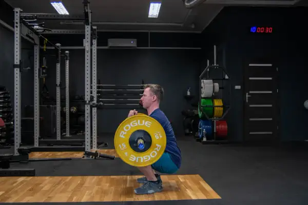 Mężczyzna wykonuje zercher squat z ciężarem w zgięciu łokci. Siłownia z wolnymi ciężarami i sprzętem do treningu siłowego.