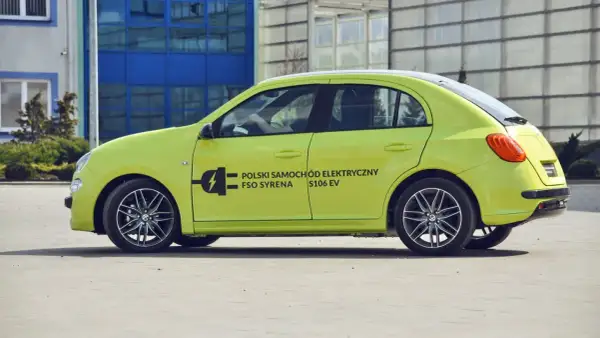 Jasnozielony FSO Syrena S106 EV, jeden z polskich samochodów elektrycznych, parkuje przed nowoczesnym budynkiem.