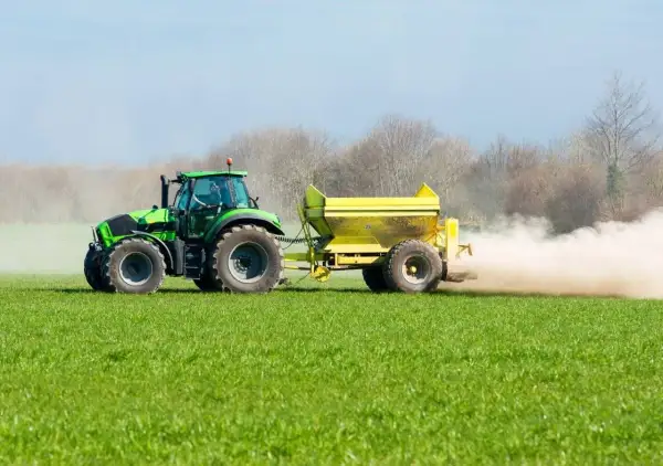 Tractor verde esparciendo cal viva en la huerta, creando una nube de polvo sobre el campo verde.