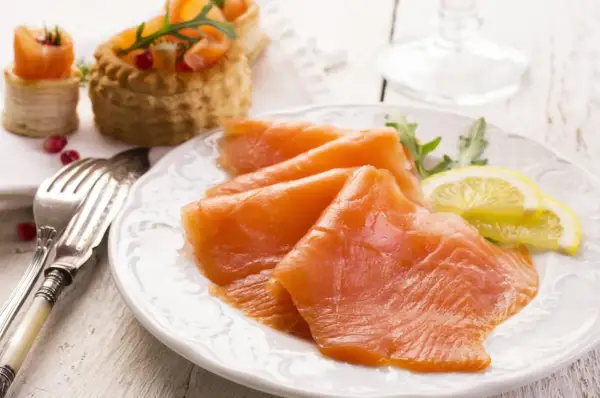 Tranches de saumon fumé, garnies de roquette et de citron, sur une assiette blanche. Des bouchées feuilletées au saumon sont visibles en arrière-plan.