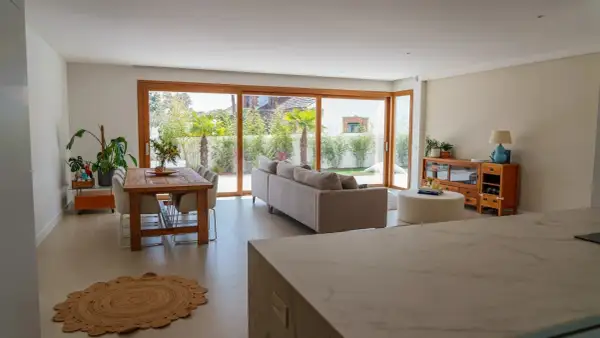 Salón moderno con comedor y sofá, destacando una pared prefabricada interior que se integra con grandes ventanales al jardín.