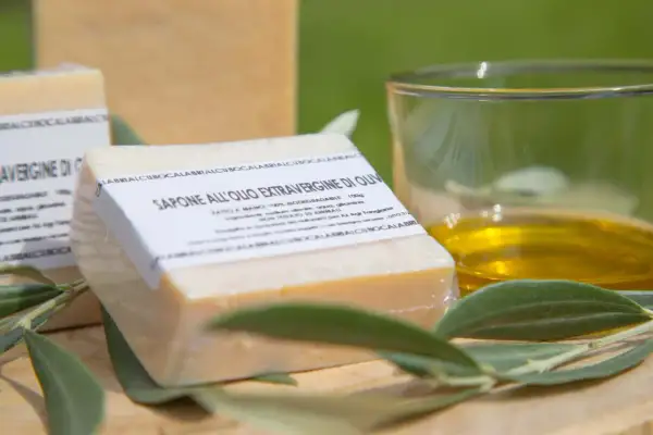 Sapone all'olio extravergine di oliva, artigianale e biodegradabile, accanto a un bicchiere d'olio e foglie d'ulivo.