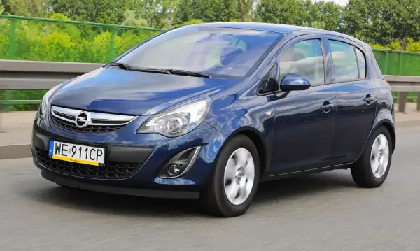 Granatowy Opel Corsa D jedzie po drodze, odbijając niebo i chmury.