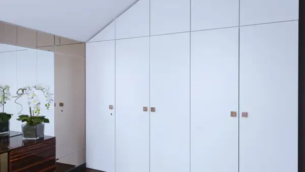Armario empotrado con puertas blancas y tiradores cuadrados. Reflejos de orquídeas y un mueble de madera oscura en el espejo. Ideal para cambiar puertas armario empotrado.