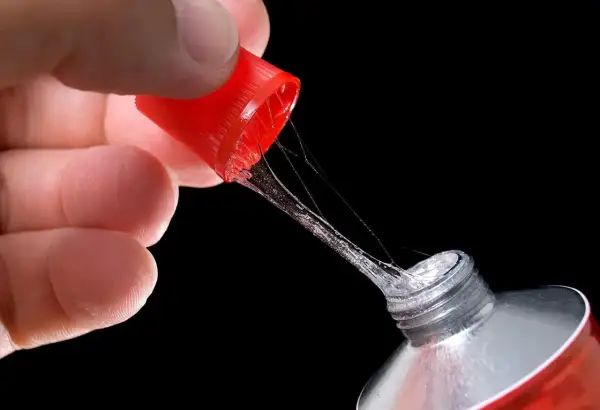 Main d'une personne tenant le bouchon rouge d'un tube de colle super glue, avec des fils de colle s'étirant. Attention, colle super glue sur les doigts dangereux.