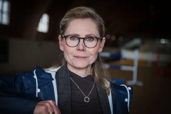 Anna Schreil med glasögon och en blå jacka, står i en ridhall.