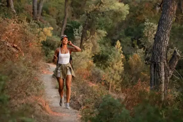 Uśmiechnięta kobieta w czapce i krótkich spodenkach uprawia power walking leśną ścieżką, słuchając muzyki.