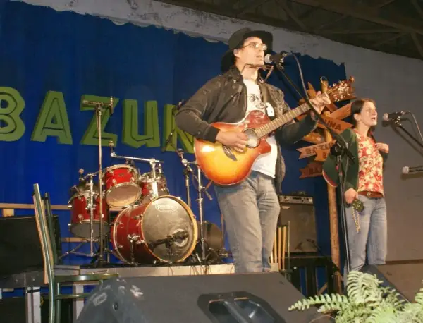 Rafał Chojnacki gra na gitarze i harmonijce ustnej, obok niego śpiewa kobieta. W tle zespół muzyczny i niebieska kurtyna z napisem "BAZUNA".