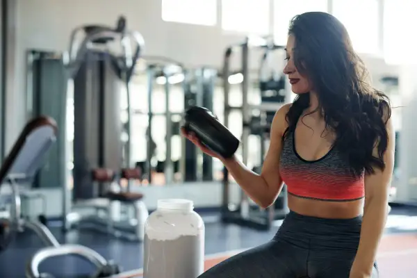 Frau im Fitnessstudio hält einen Shaker, daneben ein großer Behälter mit Pulver. Perfekt für den **proteinshake vor oder nach dem training**.