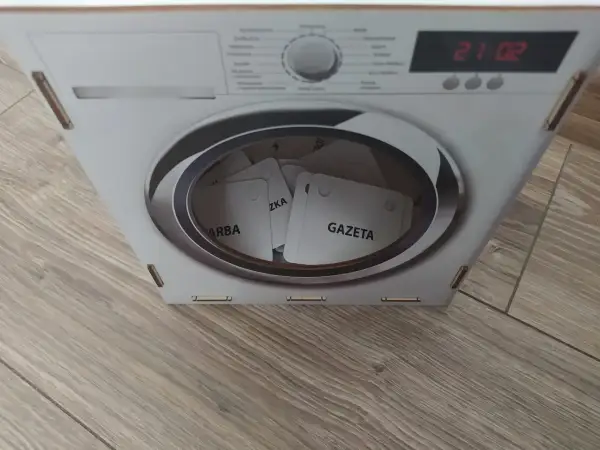 Kartonowa pralka z bębnem pełnym kart z napisami "GAZETA", "ZKA", "ARBA".