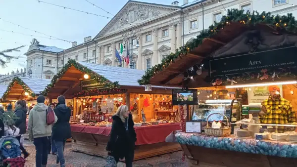 Mercatini di Natale Aosta: bancarelle addobbate con luci e ghirlande, prodotti tipici e gente che passeggia. Atmosfera magica.