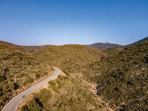 Vue aérienne d'une route sinueuse traversant des collines verdoyantes, menant vers le col du Testanier. Deux cyclistes s'engagent sur la pente.