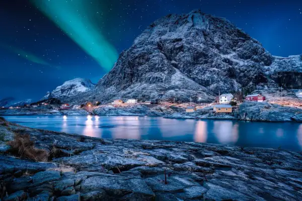 Grüne Bänder der Aurora Borealis tanzen über einem verschneiten Berg, der eine kleine Küstensiedlung mit roten und gelben Häusern beleuchtet.