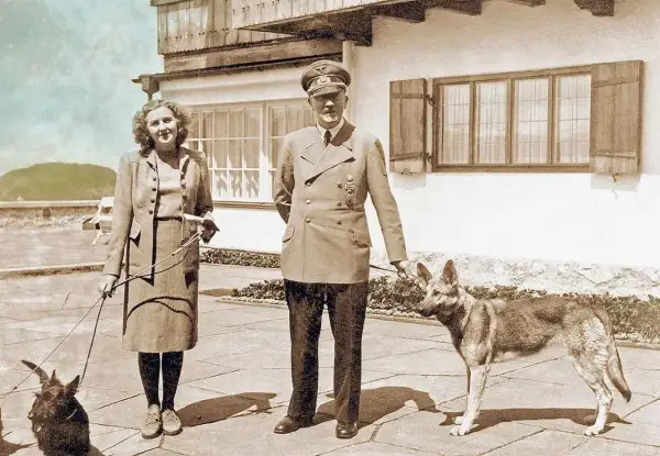 Eva Braun, hitlers fru, står med en hund. Bredvid henne står en man i uniform med en annan hund.
