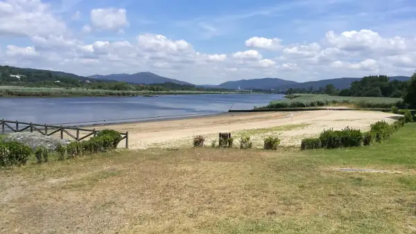 La playa fluvial Vilarello, con arena dorada y aguas tranquilas, se extiende ante colinas verdes bajo un cielo azul con nubes blancas.