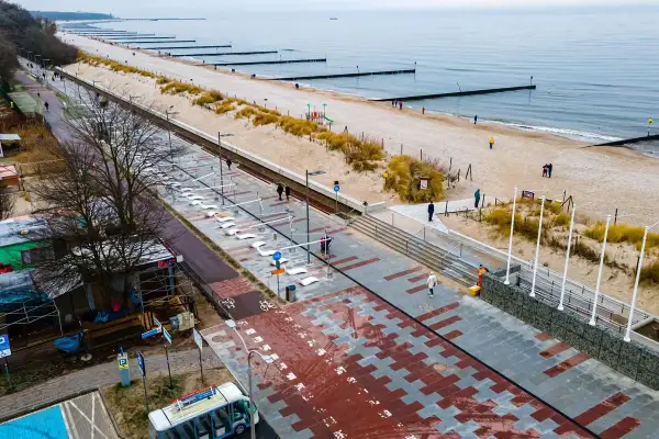 Nowoczesna promenada nad morzem, z piaskiem, wydmami i łagodnymi falami. Ludzie spacerują, ciesząc się chłodnym dniem.