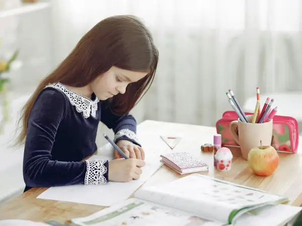 Niña concentrada en su dictado 2 primaria, rodeada de útiles escolares, un libro abierto y una manzana.