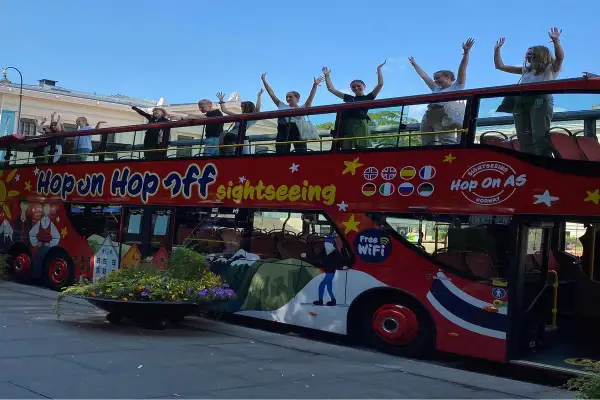 Glada resenärer vinkar från övervåningen på en röd dubbeldäckare med texten "Hop on Hop off sightseeing".